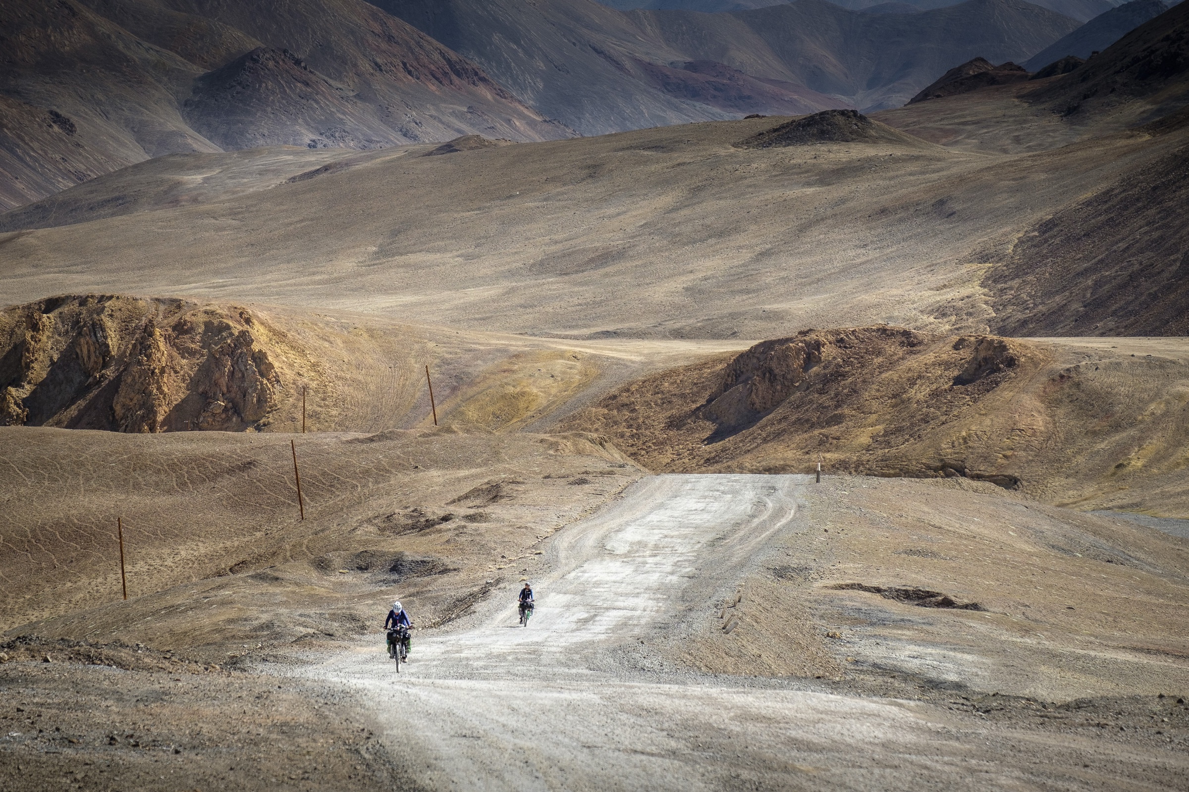 Cyclists following a winding road through a barren mountain valley in Central Asia.