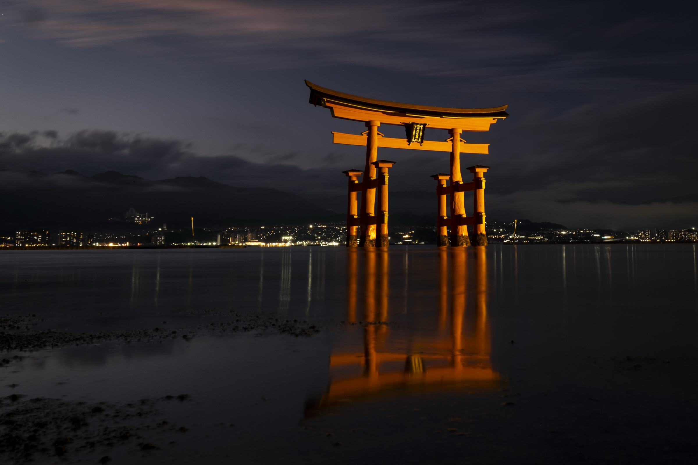 An illuminated orange torii gate reflected in still water at dusk in Japan.