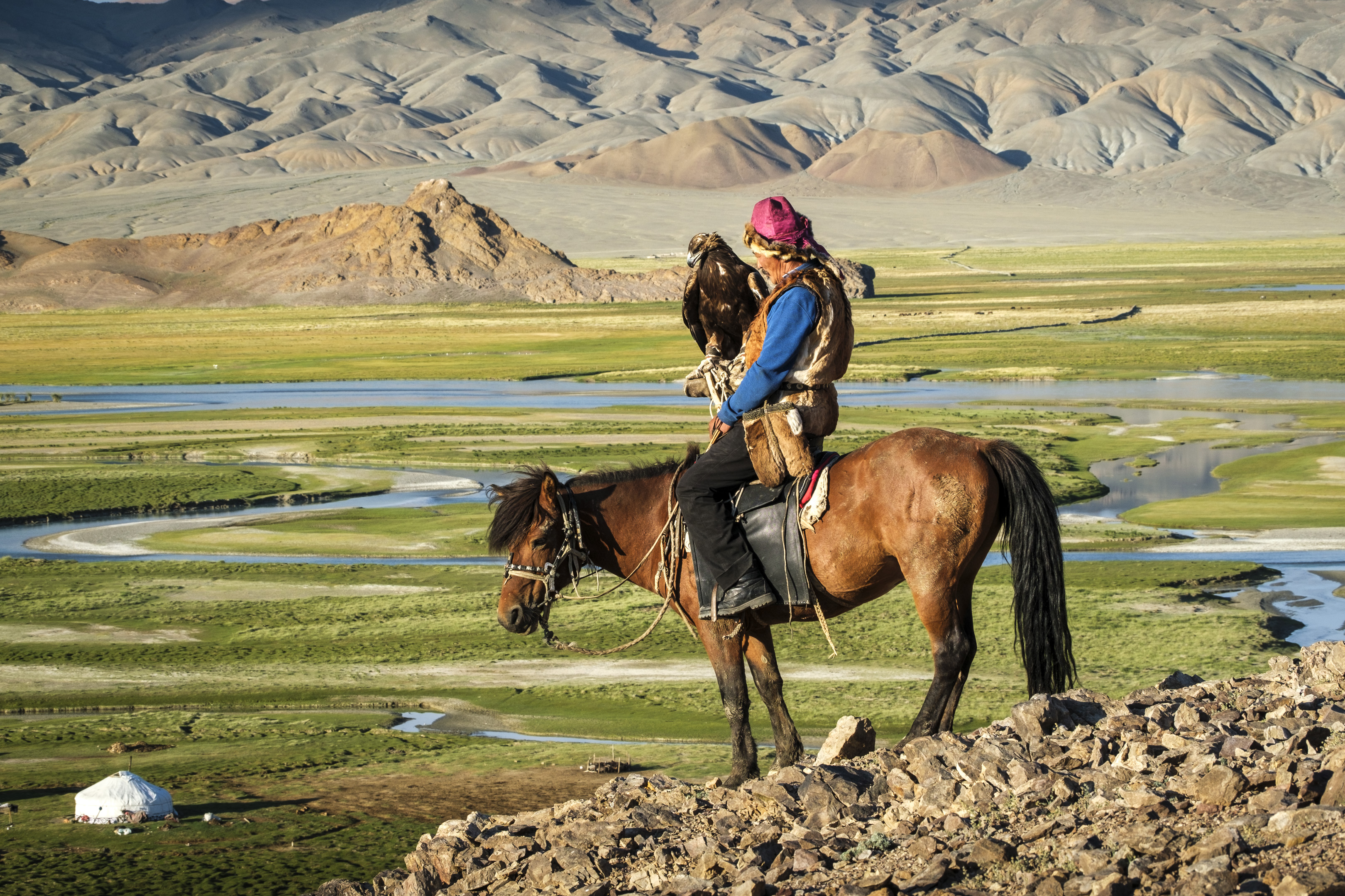 An eagle hunter on horseback beside a river in a mountain valley in Mongolia.